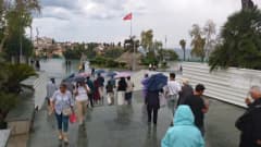 Antalya Cumhuriyet Meydanı’nda yağmur altında şemsiyelerini açmış turistler ve yağmur bulutlarıyla Akdeniz manzarası