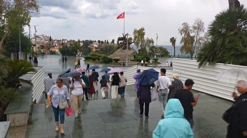 Antalya Cumhuriyet Meydanı’nda yağmur altında şemsiyelerini açmış turistler ve yağmur bulutlarıyla Akdeniz manzarası